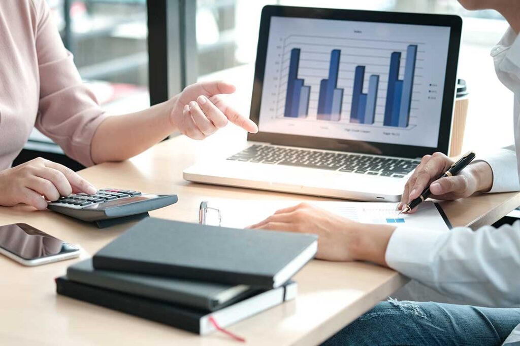 A person reviewing their checkbook and mobile banking app at a kitchen table with bills and a calculator.